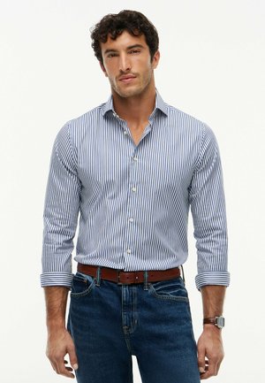 Mann trägt ein blau-weiß gestreiftes Hemd mit hochgekrempelten Ärmeln, dunkelblaue Jeans, braunen Gürtel und Armbanduhr, steht frontal.