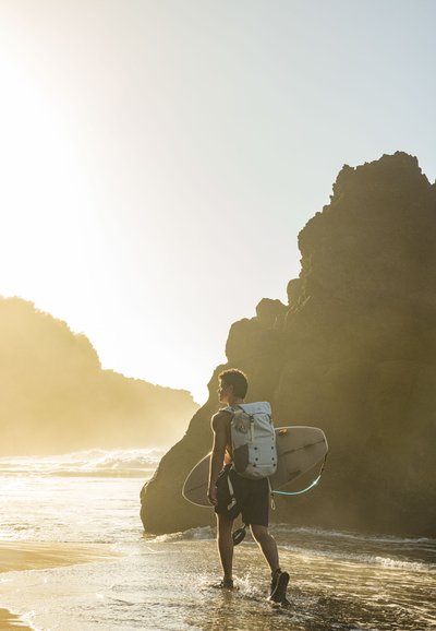Surfista con una mochila gris claro y una tabla de surf, caminando sobre arena mojada cerca de formaciones rocosas oscuras, con luz solar realzando la escena.