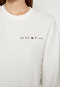 Sweat-shirt blanc avec inscription GANT 1949 et un petit logo en forme de bouclier avec des rayures rouges, blanches et bleues et des étoiles sur la poitrine.