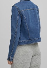 Džínová bunda v modré barvě s klasickým límcem, dlouhými rukávy a rovný střih. Obsahuje prošívané švy a knoflíkové manžety.