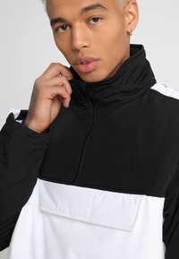 Svart og hvit pullover-jakke med høyt krage, frontlomme og strukturert stoff. Har også en frontglidelås og fargeblokkdesign.