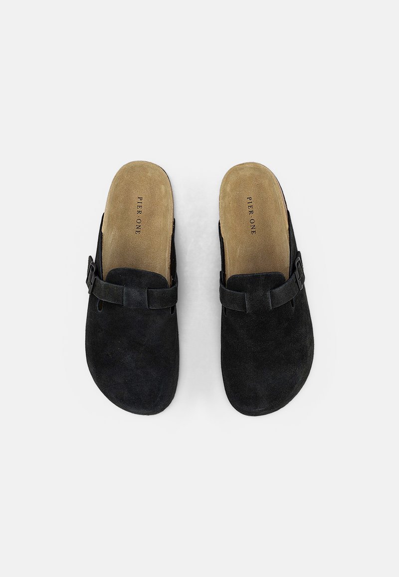 Chaussures noires en daim à enfiler avec une forme arrondie, dotées d'une semelle plate et d'une sangle avant avec détail de boucle. L'intérieur doublé affiche le nom de la marque.