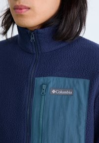 Navy fleece jacket with high collar, zip front, and contrasting teal chest pocket featuring a zipper and logo patch. Soft texture.