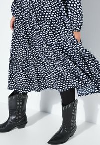 Musta mekko, jossa valkoinen sydänkuvio, kerroksittainen muotoilu ja pitkät hihat. Yhdistetty polvimittaisiin mustiin cowboy-saappaisiin, joissa hopeiset niitit.