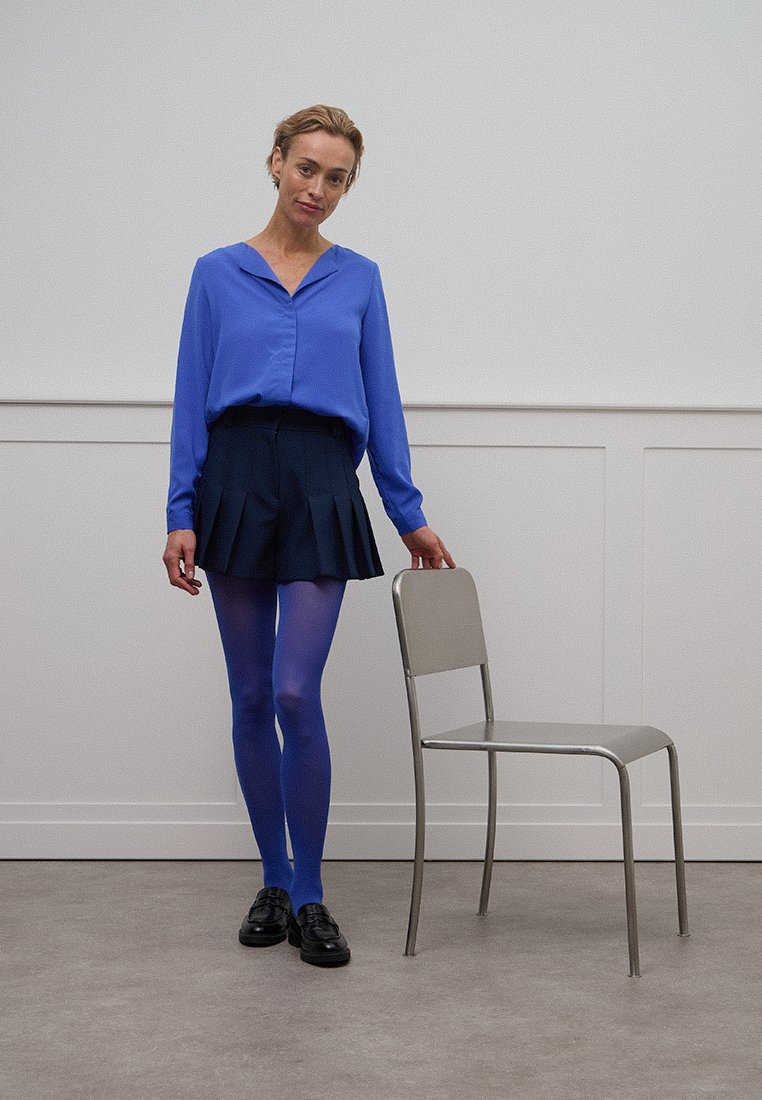 Helles blaues Blusenshirt mit Kragen, dunkler gefalteter Minirock und blauen Strumpfhosen. Schwarze Schuhe und ein metallisch grauer Stuhl vor schlichem Hintergrund.