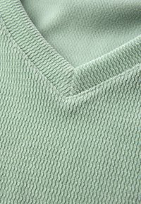 Groene, gestructureerde stofstaal met een V-hals ontwerp. Het materiaal heeft verticale ribbels en een over het algemeen gladde afwerking.