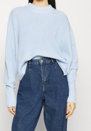Pull en maille bleu clair avec un col montant et des épaules tombantes, associé à un jean en denim taille haute avec une fermeture à bouton.
