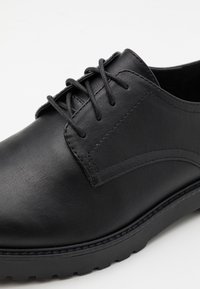 Chaussure habillée noire en cuir avec lacets, détails cousus et semelle épaisse en caoutchouc, vue en angle sur fond blanc.