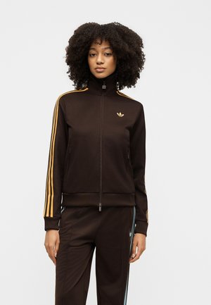 Frau mit lockigem Haar, die einen braunen Adidas-Trainingsanzug mit orangenen Streifen an den Ärmeln und hellblauen Streifen an der Hose trägt und vor weißem Hintergrund steht.