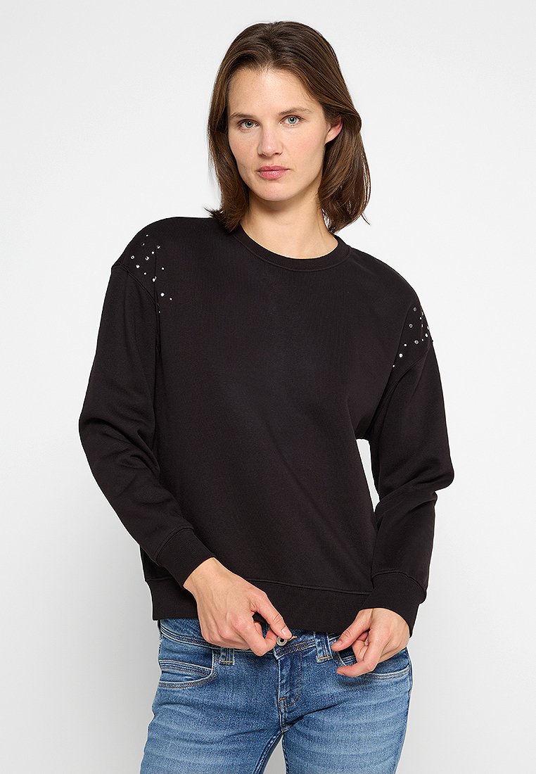 Sudadera negra de tejido acanalado, con acentos de tachuelas en los hombros, cuello redondo y puños acanalados, combinada con jeans azules.