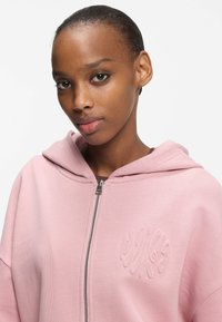 Light pink zip-up hoodie with a drawstring hood, featuring an embroidered design on the chest and a soft, textured fabric.