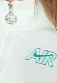 Myntgrön zip-up jacka med ett texturerat tyg, som har en broderad "AIR"-logotyp i teal och silverfärgad hårdvara för dragkedjan.