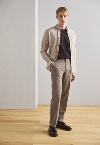 O cămașă button-up bej deschis peste un tricou negru, asortată cu pantaloni gri deschis și pantofi negri, pe un fundal neutru.