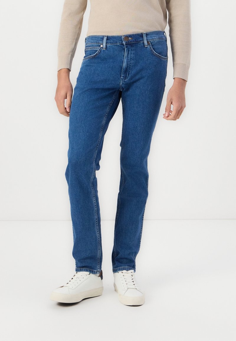 Jean en denim bleu à coupe droite, style cinq poches, fermeture par bouton, associé à des baskets blanches.
