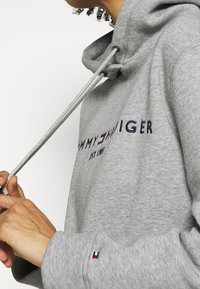Person som bär en grå Tommy Hilfiger-hoodie, justerar snöret; logotyp och liten flaggemblem syns på bröstet och ärmen.