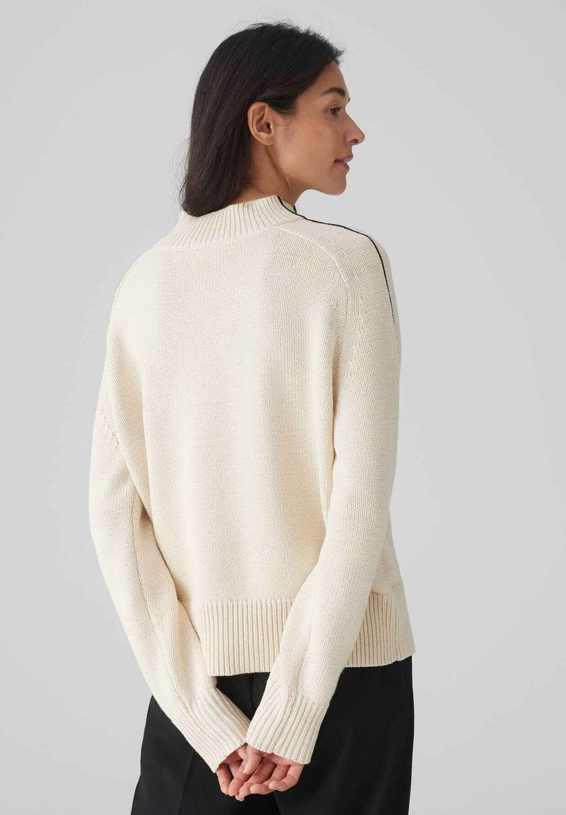 Cremfarbener Strickpullover mit gerippten Bündchen und Saum, der einen hohen Kragen und kontrastierende schwarze Streifen an den Schultern aufweist.