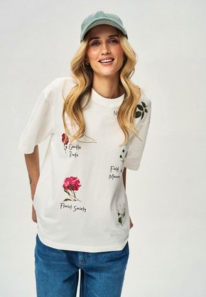 Uśmiechnięta kobieta o długich blond włosach nosi zieloną czapkę, białą koszulkę z nadrukiem kwiatowym i napisem oraz niebieskie dżinsy na jednolitym tle.