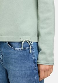 Hellgrüner Baumwollpullover mit Kordelzug am Saum, kombiniert mit blauen Jeans. Die Textur ist glatt, mit enganliegenden Bündchen.