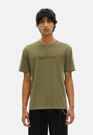 Jongeman met donker haar die een olijfgroen T-shirt draagt met vervaagde zwarte tekst en zwarte broek met een zilveren kettingaccessoire.