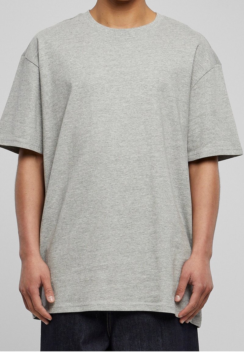Personne portant un t-shirt ample à manches courtes gris clair uni et un pantalon foncé, debout devant un fond neutre.