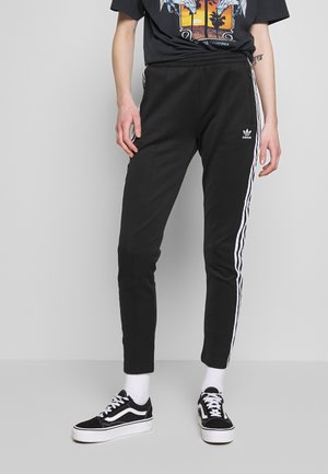Personne portant un pantalon de survêtement Adidas noir à bandes blanches, associé à des baskets noires et blanches et un t-shirt noir graphique.