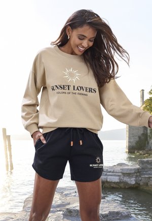 Beige Sweatshirt mit Sonnenmotiv und schwarzer Schrift, kombiniert mit schwarzen Shorts mit elastischem Bund und hellbraunen Kordelzug-Details.