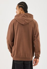 Felpa con cappuccio marrone, dal taglio rilassato, in tessuto testurizzato e con polsini a coste. Cuciture visibili lungo le spalle e la schiena.