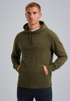Homme aux cheveux courts portant un hoodie vert olive avec une poche devant et un petit logo sur la poitrine, se tenant devant un fond gris uni.