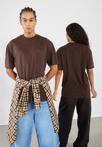 Camiseta marrón de manga corta con bolsillo en el pecho; combinada con una camisa de franela a cuadros atada alrededor de la cintura; el modelo lleva puestos unos jeans de pierna ancha en azul claro.