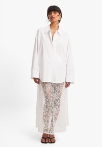 Camisa blanca de botones con mangas largas, combinada con pantalones de encaje blanco transparente que presentan un patrón floral y una caída larga en la parte posterior. Sandalias negras.