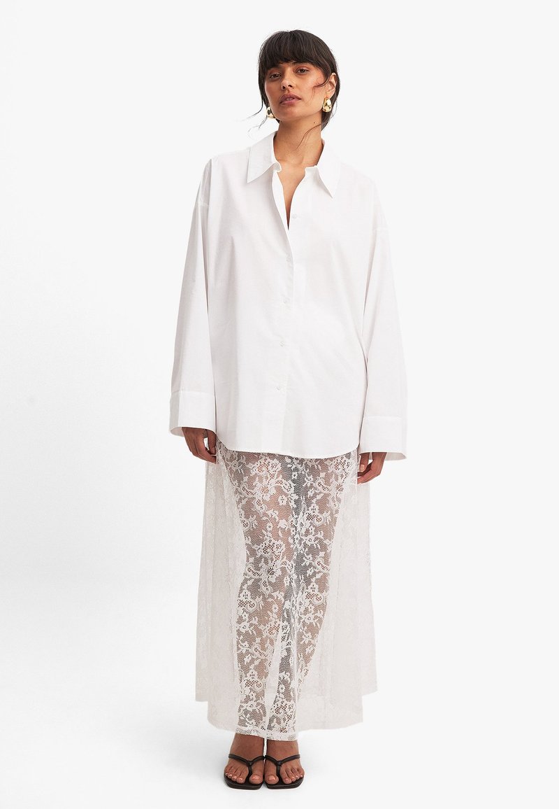 Camisa blanca de botones con mangas largas, combinada con pantalones de encaje blanco transparente que presentan un patrón floral y una caída larga en la parte posterior. Sandalias negras.