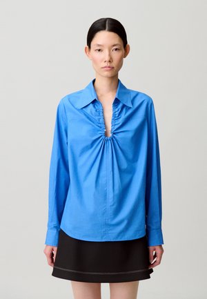 Camicia blu a maniche lunghe con scollo arricciato, caratterizzata da un dettaglio con bottone. Abbinata a una gonna nera con cuciture a contrasto.