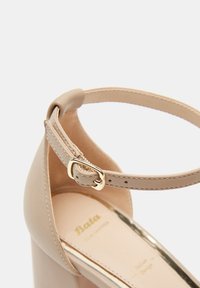 Zapato de tacón alto beis con acabado de cuero suave, correa para el tobillo y hebilla dorada, que presenta un borde metálico en la suela.