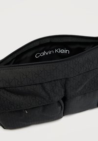 Μαύρο τσαντάκι Calvin Klein με κλείσιμο με φερμουάρ. Διαθέτει υφή υφάσματος με διακριτικό μοτίβο. Εσωτερική επένδυση με το λογότυπο της μάρκας.