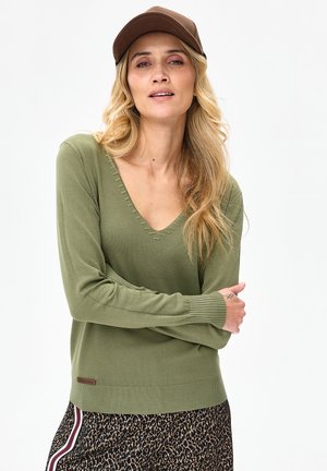 Kobieta z długimi blond włosami, ubrana w brązową czapkę, oliwkowozielony sweter z dekoltem w serek oraz spódnicę w panterkę z bocznymi paskami, stojąca z założonymi ramionami.