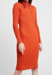 Ribbad orange stickad klänning med långa ärmar, hög krage och vågig nederkant. Har en strukturerad kabeldesign längs fronten.