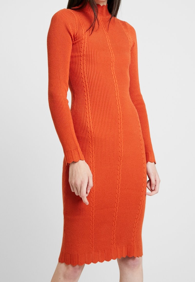 Ribbad orange stickad klänning med långa ärmar, hög krage och vågig nederkant. Har en strukturerad kabeldesign längs fronten.