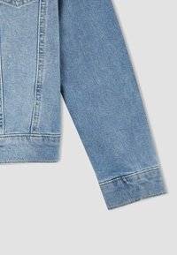 Una chaqueta de mezclilla azul claro presenta un cuello estándar, mangas largas y costuras visibles a lo largo de las costuras. La tela tiene un acabado texturizado.