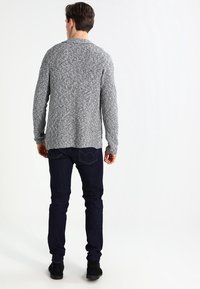 Maglione grigio mélange a maglia, collo a giro, maniche lunghe, orlo leggermente più lungo sul retro, abbinato a jeans in denim scuri e stivali neri.