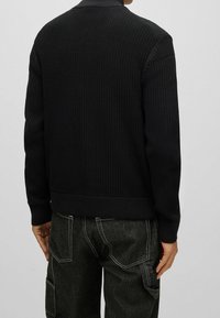 Maglione nero a maglia con texture a coste, dotato di un collo alto e polsini aderenti, abbinato a pantaloni cargo scuri.