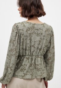 Blusa verde oliva con sovrapposizione di pizzo floreale, maniche lunghe e vita arricciata, caratterizzata da un colletto rotondo e una trama trasparente.