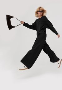 Czarny, oversize sweter i spodnie z szerokimi nogawkami o gładkiej fakturze. Białe sneakersy z brązową podeszwą. Trzymanie ciemnobrązowej torby z paskiem.