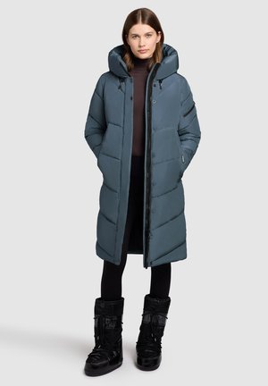 Lang, gevoerde pufferjas in leisteenblauw met een grote capuchon, frontrits, drukknoopsluiting en twee zijzakken. Draagt zwarte sneeuwlaarzen.