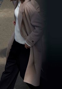 Dlouhý béžový vlněný kabát se strukturovaným designem, vybavený vysokým límcem, knoflíkovými detaily a volným střihem, doplněný bílým žebrovaným svetrem a černými kalhotami.