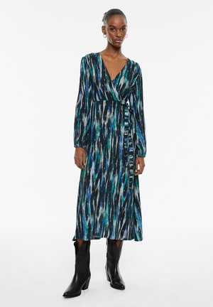 Mujer vestida con un vestido envolvente de manga larga, de patrón azul y verde, combinado con botas negras hasta la mitad de la pantorrilla, de pie frente a un fondo blanco.