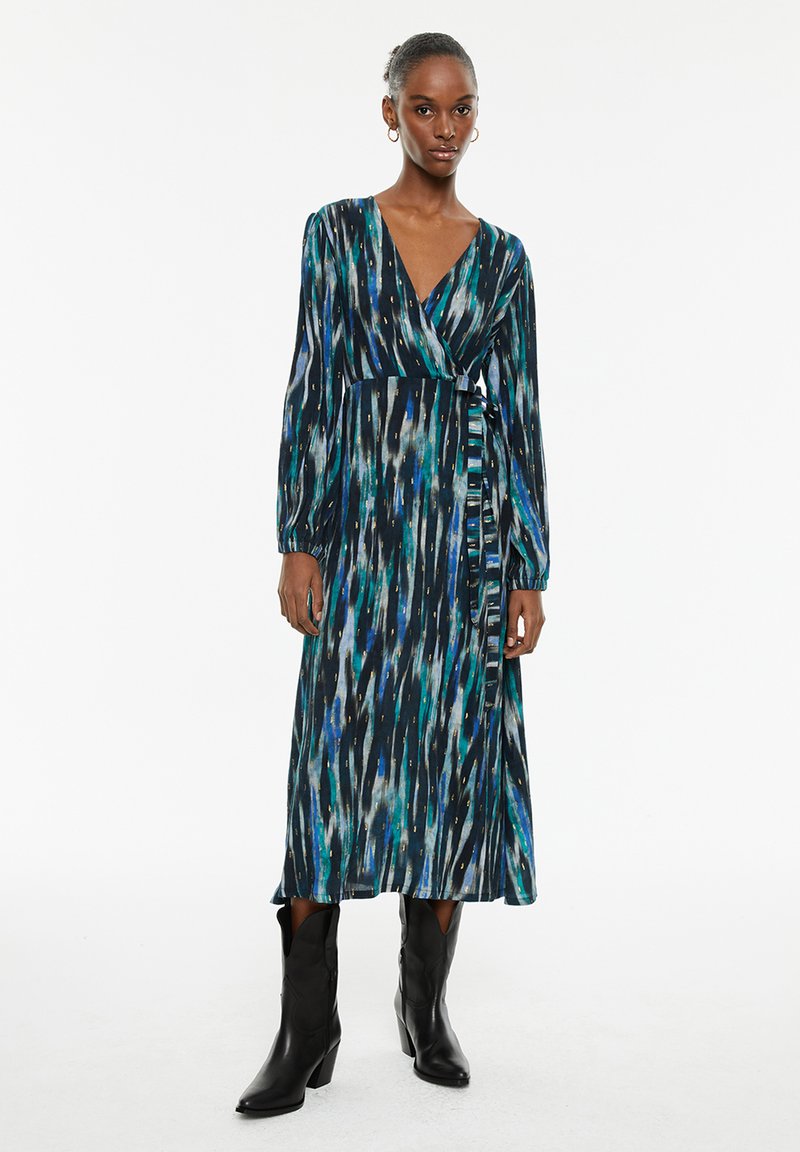 Mujer vestida con un vestido envolvente de manga larga, de patrón azul y verde, combinado con botas negras hasta la mitad de la pantorrilla, de pie frente a un fondo blanco.