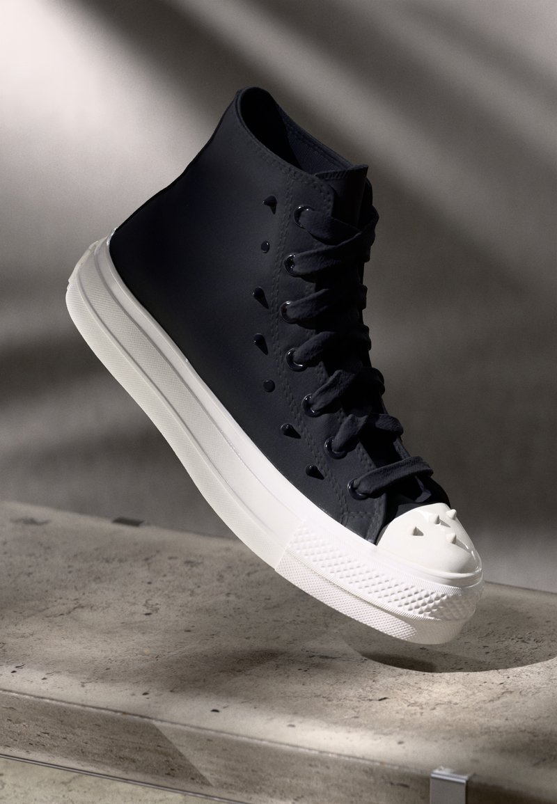 Baskets montantes noires avec semelle blanche et embout pointu blanc reposant sur une surface en béton gris avec une ombre douce en arrière-plan.