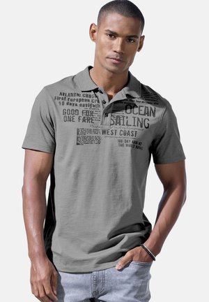 Mann in grauem Poloshirt mit schwarzem Segelmotiv-Text, hellen Jeans und schwarzem Armband steht vor hellem, einfarbigem Hintergrund.