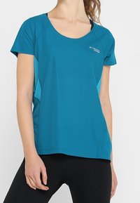 Kvinna som bär en turkos Columbia sporttröja och svarta leggings, visad från nacken till höfterna mot en enkelfärgad ljus bakgrund.