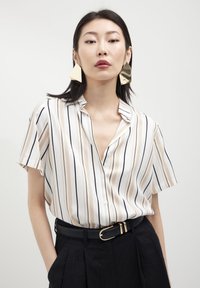 Camicia a maniche corte con colletto, a righe verticali beige e nere su uno sfondo bianco, abbinata a pantaloni neri a vita alta e una cintura nera.